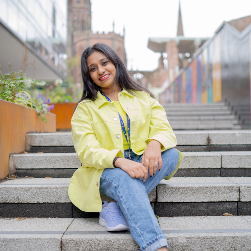 Panchami, a young woman with dark long hair, sitting on stair next to the hub and smiling at the camera.
