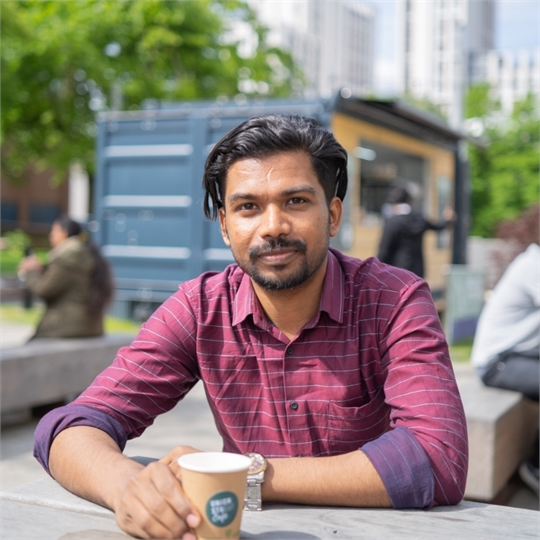 Akhil, a young man with medium length, dark hair and a beard, sitting in front of the Union Street Cafe and smiling at the camera.