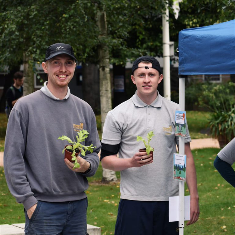 Two young male students looking at the camera smiling and holding pots with plants.