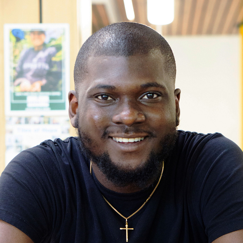 Desmond, a young black man with close cropped hair and a chinstrap beard smiling at the camera. He's wearing a black t-shirt and a gold necklace.