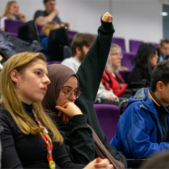 A group of students in a lecture hall, 1 student has their hand up