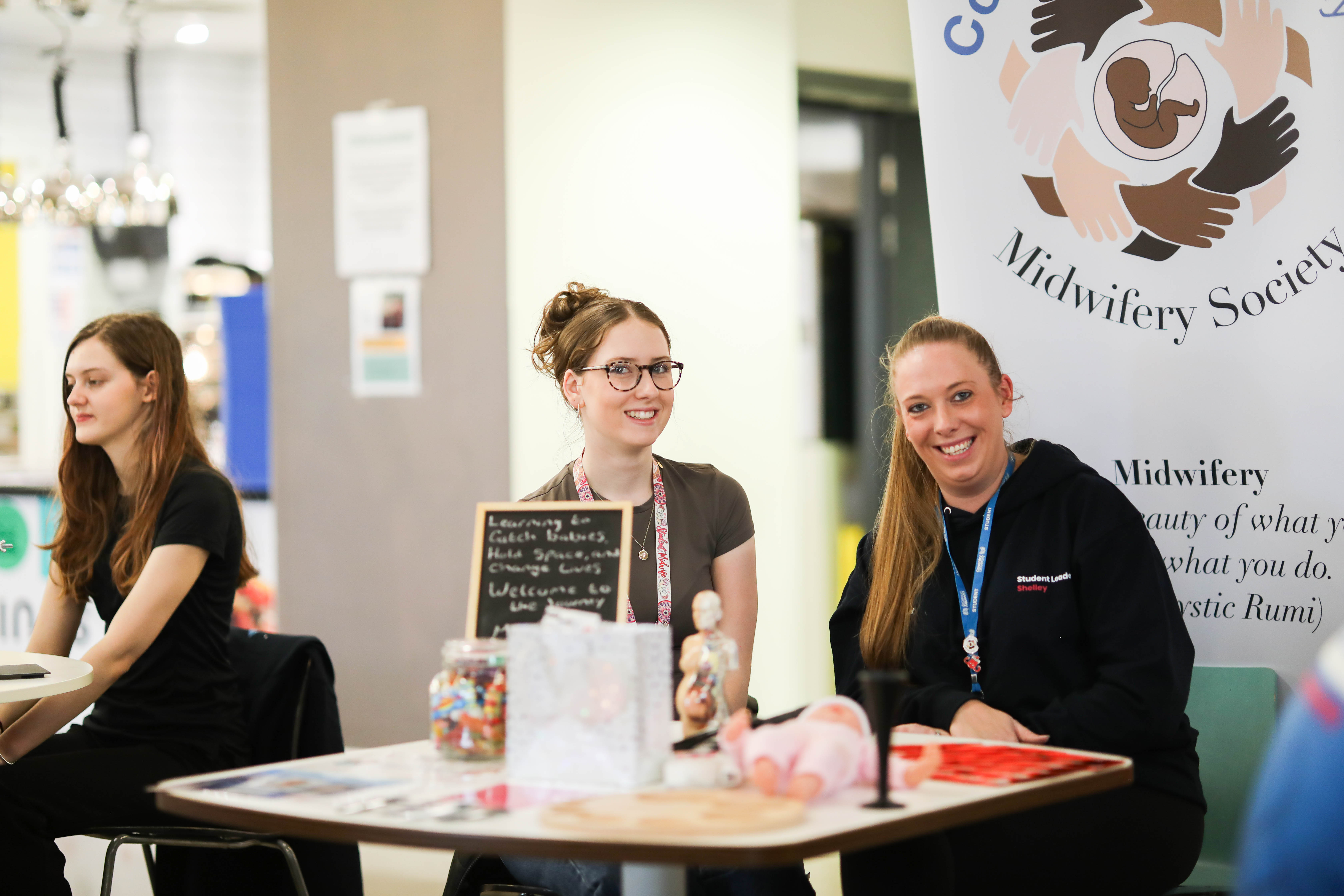 Two people sitting behind a table in front of a Midwifery Society banner.