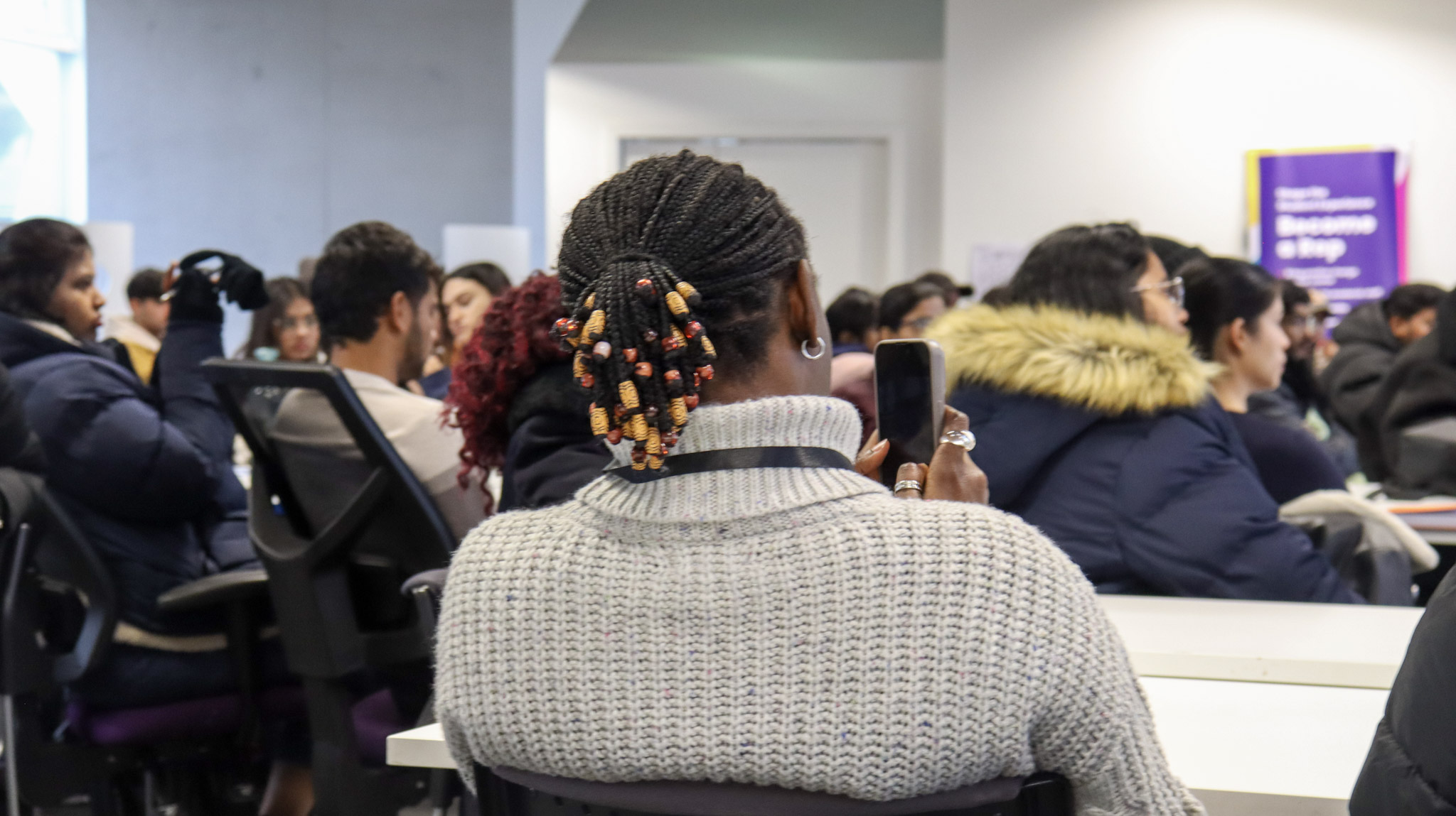 The back of a person sitting down listening to a speaker at the front of a room.
