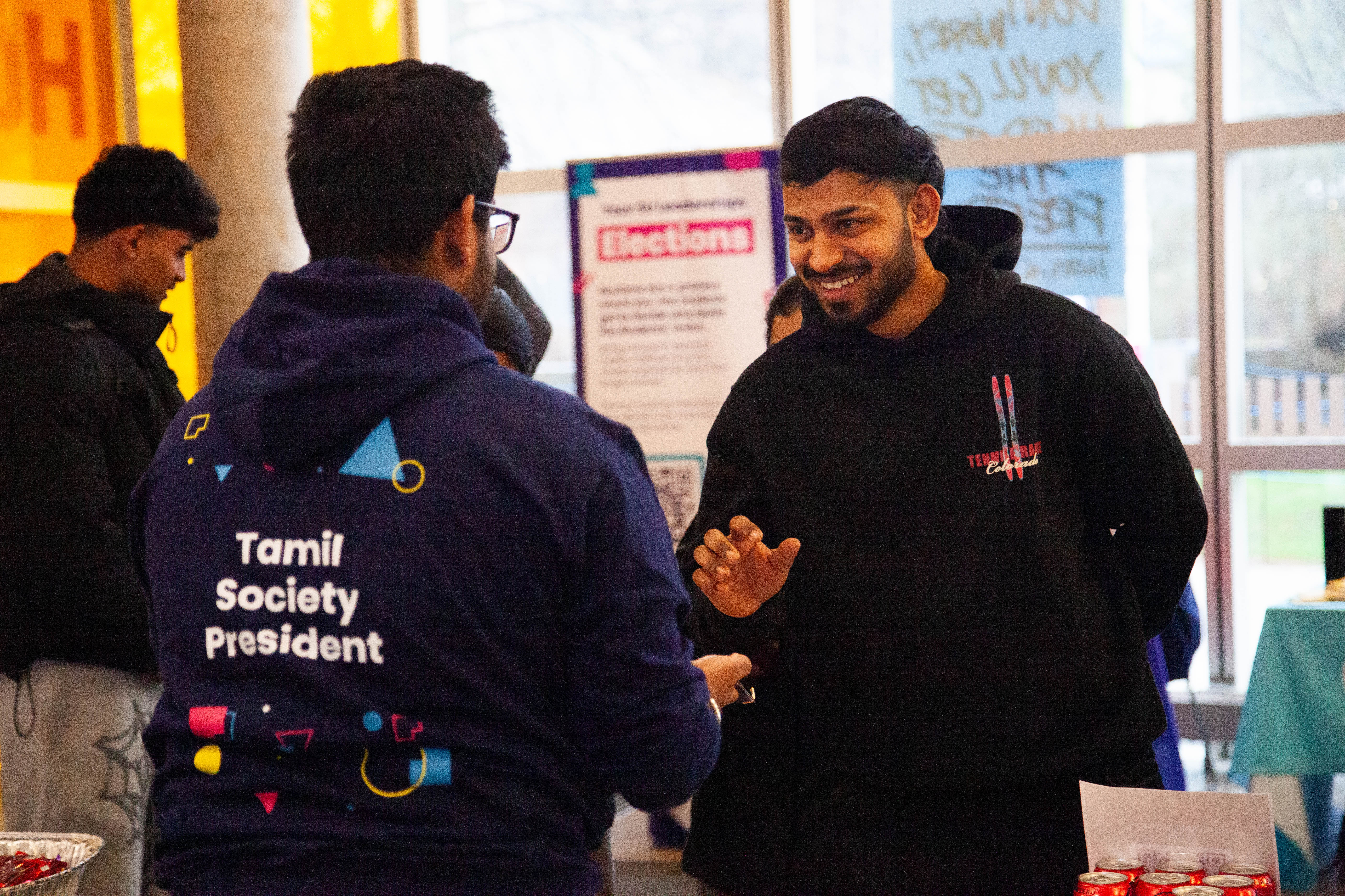 Two people standing and talking. One person wearing a SU-branded hoodie reading: 'Tamil Society President'.