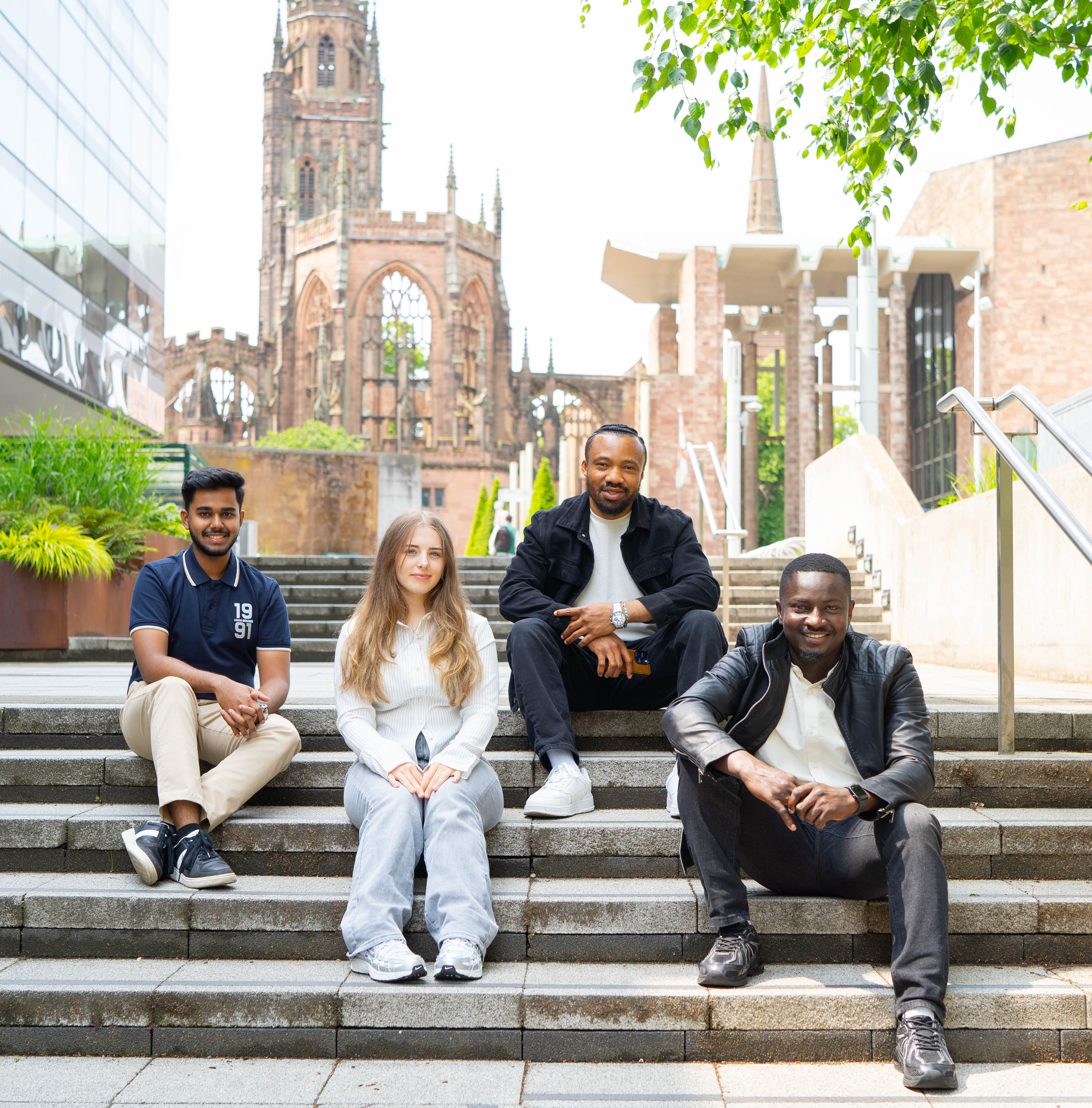 Four people sat on stairs with the Coventry Cathedral in the background.