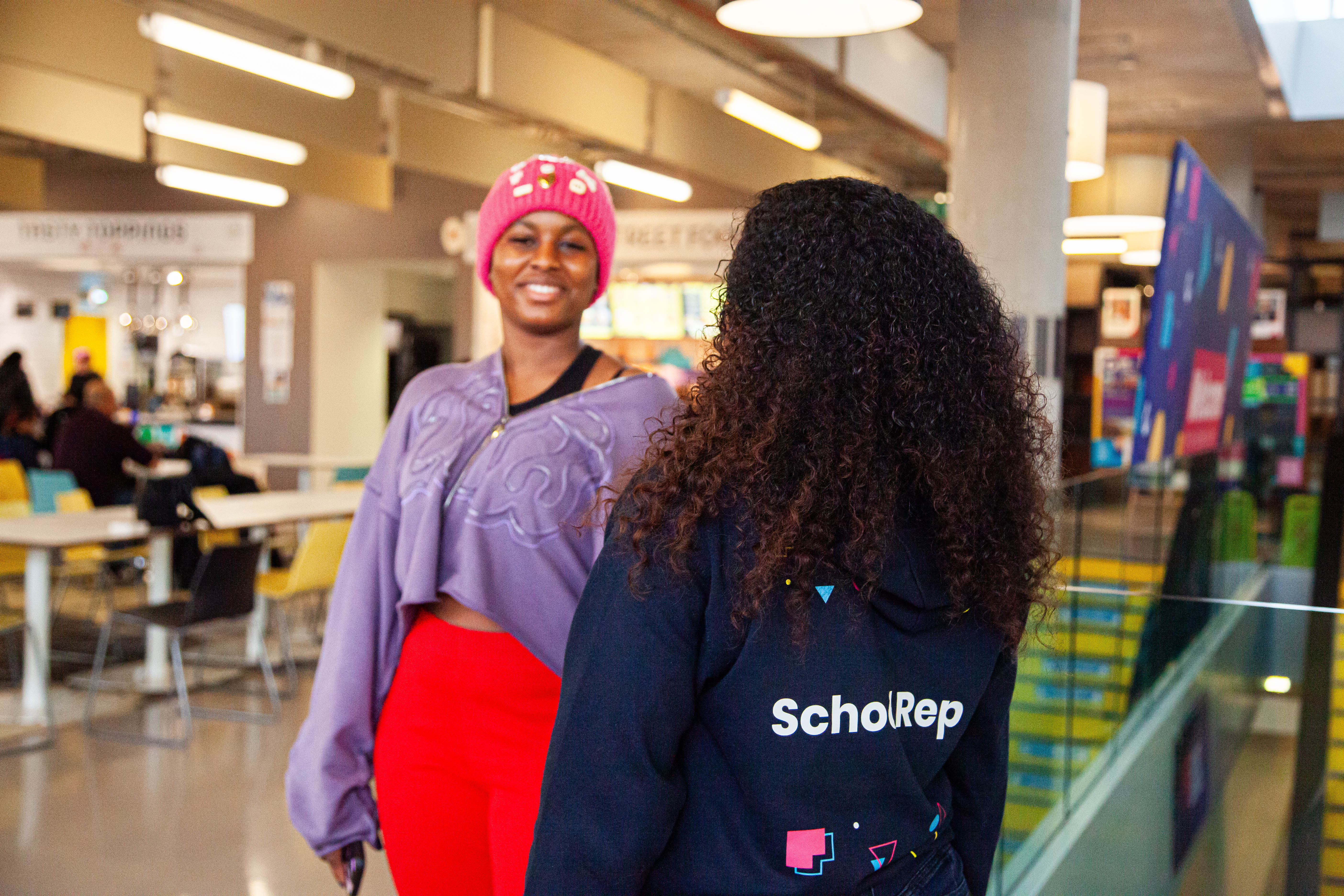 Two people standing and having a conversation, with the back of one person wearing a School Rep hoodie.