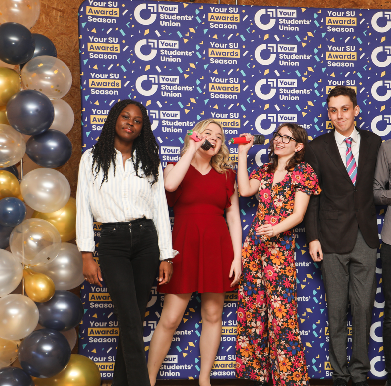 Four people stood in front of a Students' Union branded banner. The middle two people are speaking into toy microphones.